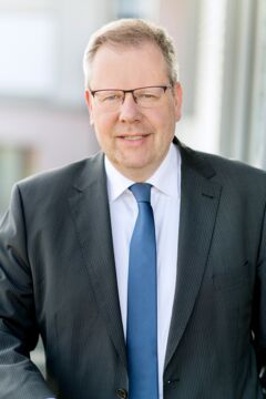 Ein Mann mit grauem Haar und Brille trägt einen dunklen Anzug und steht vor einem Fenster